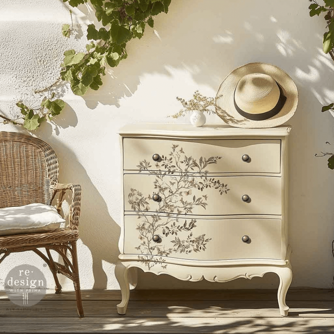 Decorative chest of drawers with floral patterns in a sunlit room with a wicker chair and straw hat.