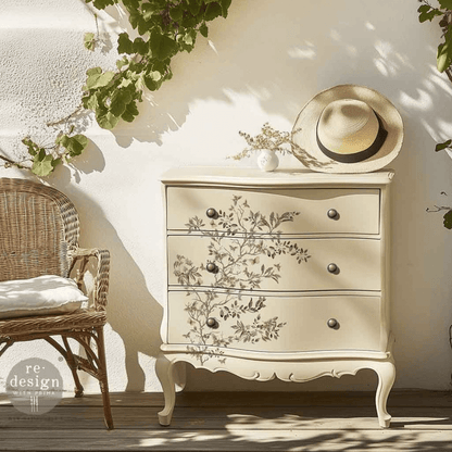 Decorative chest of drawers with floral patterns in a sunlit room with a wicker chair and straw hat.