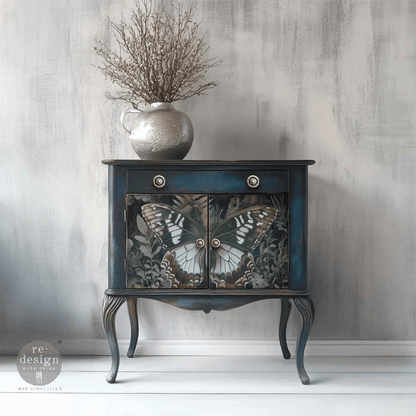 A small vintage-style side cabinet with a dark blue painted frame and butterfly artwork on the doors, styled with a silver vase of dried branches on top against a textured gray wall.
