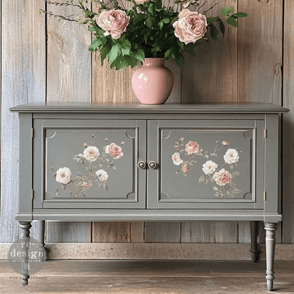 Gray cabinet with floral designs on doors, pink vase with flowers on top against a wooden wall.