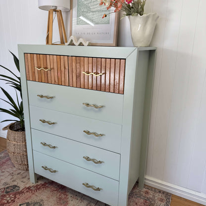A tall chest of drawers painted with Blake & Taylor Furniture paint, colour Willow styled in a home setting with a lamp, framed art, and flowers on top.