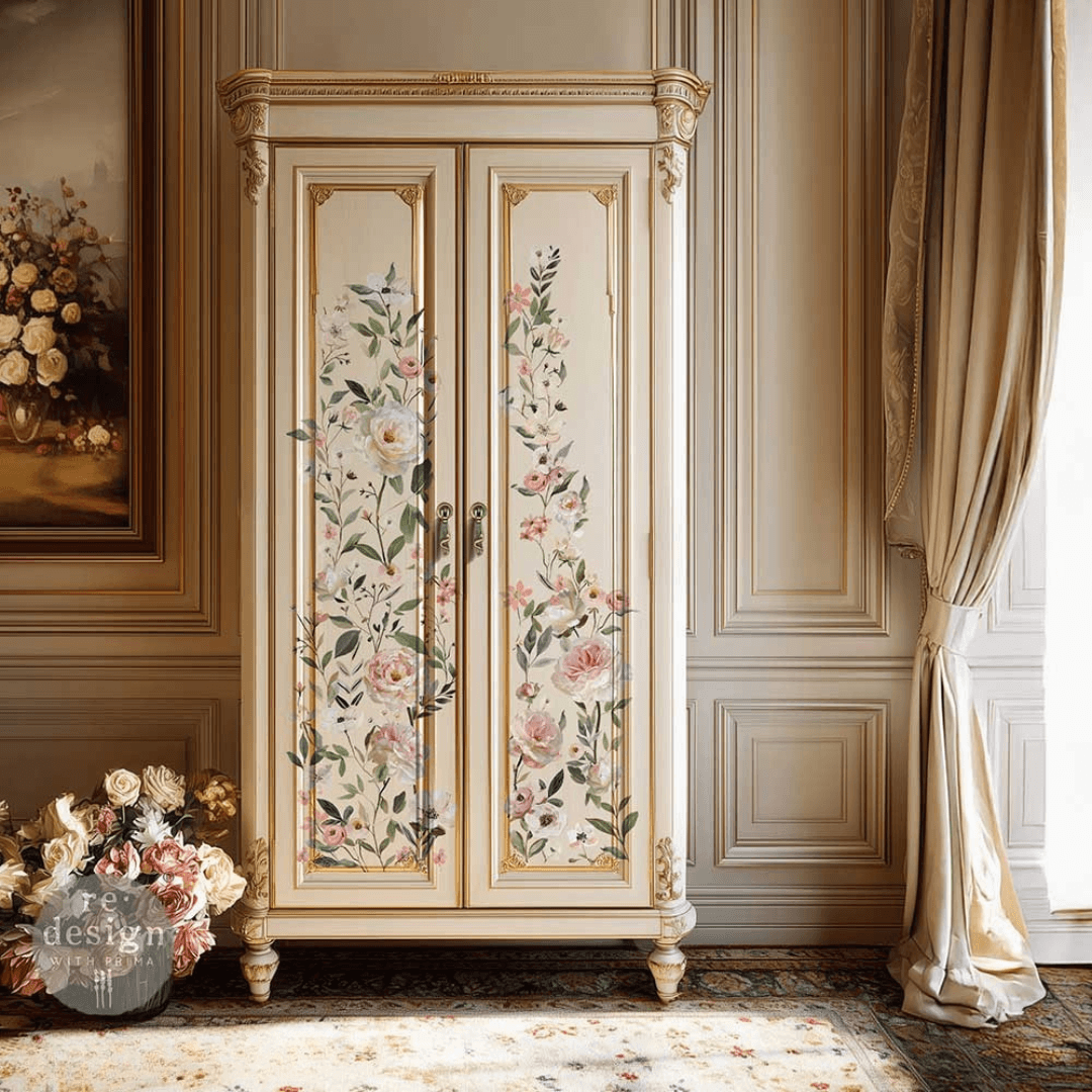 An ornate vintage-style double-door cabinet or wardrobe in a warm cream/beige finish, decorated with painted floral motifs on both doors. It stands in a classical interior with paneled walls, draped curtains, a patterned rug, and a framed floral painting.
