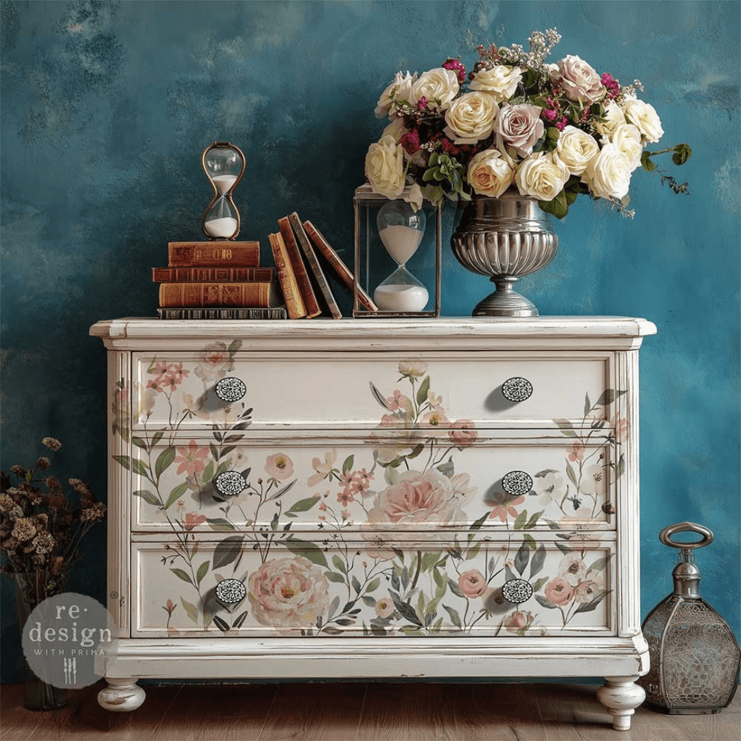 A vintage-style three-drawer dresser with a distressed cream finish and a delicate pink and green floral print, styled with books, an hourglass, and a large bouquet of roses against a textured teal wall.