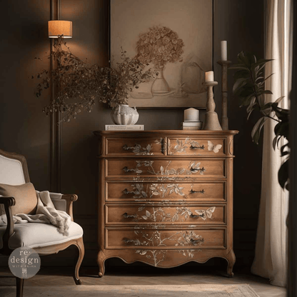 A warm brown wooden chest of drawers with delicate white floral inlay details, styled in a cozy, dark-walled living room next to a white armchair and a floor lamp.