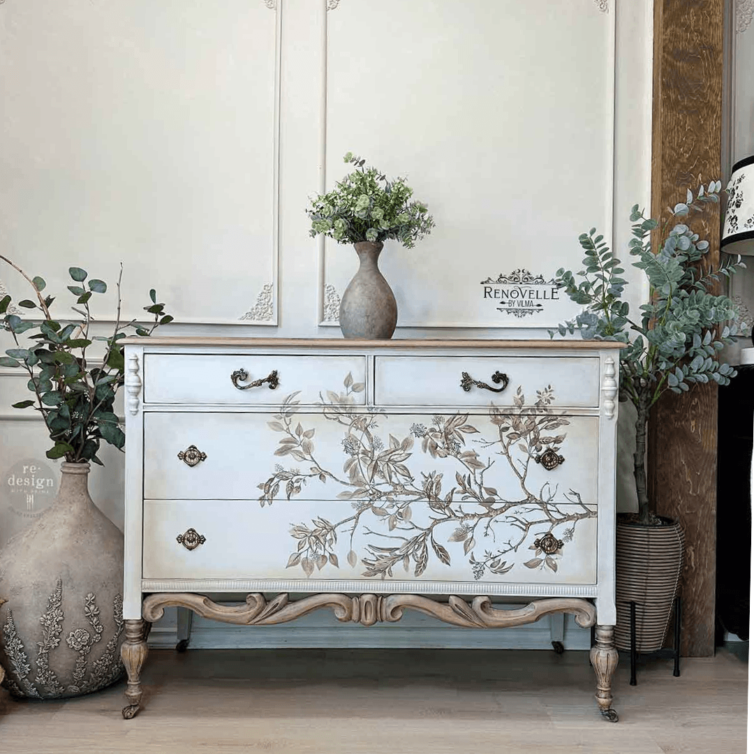 A vintage-style white chest of drawers with carved legs and floral branch illustrations on the front, styled in a room setting with decorative vases and plants.