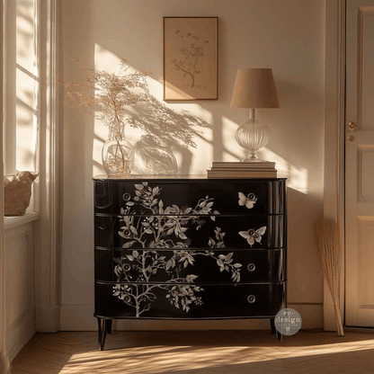 A glossy black chest of drawers with white floral and butterfly decals, styled in a sunlit room with a lamp, vase, and framed art on top.