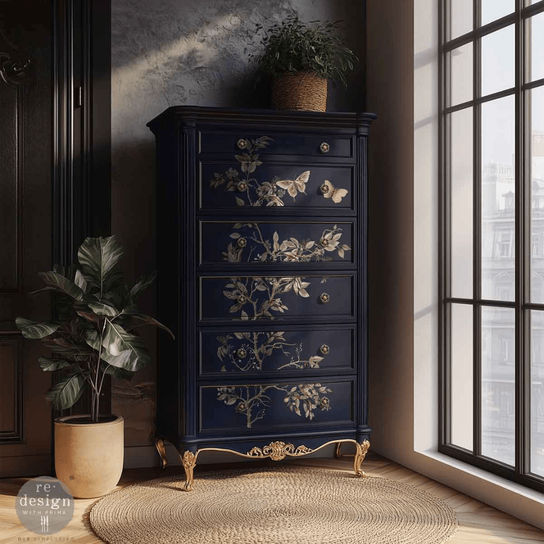 A tall dark navy blue five-drawer chest of drawers with ornate gold floral and butterfly motifs on the drawer fronts, styled in a room next to a window with potted plants.