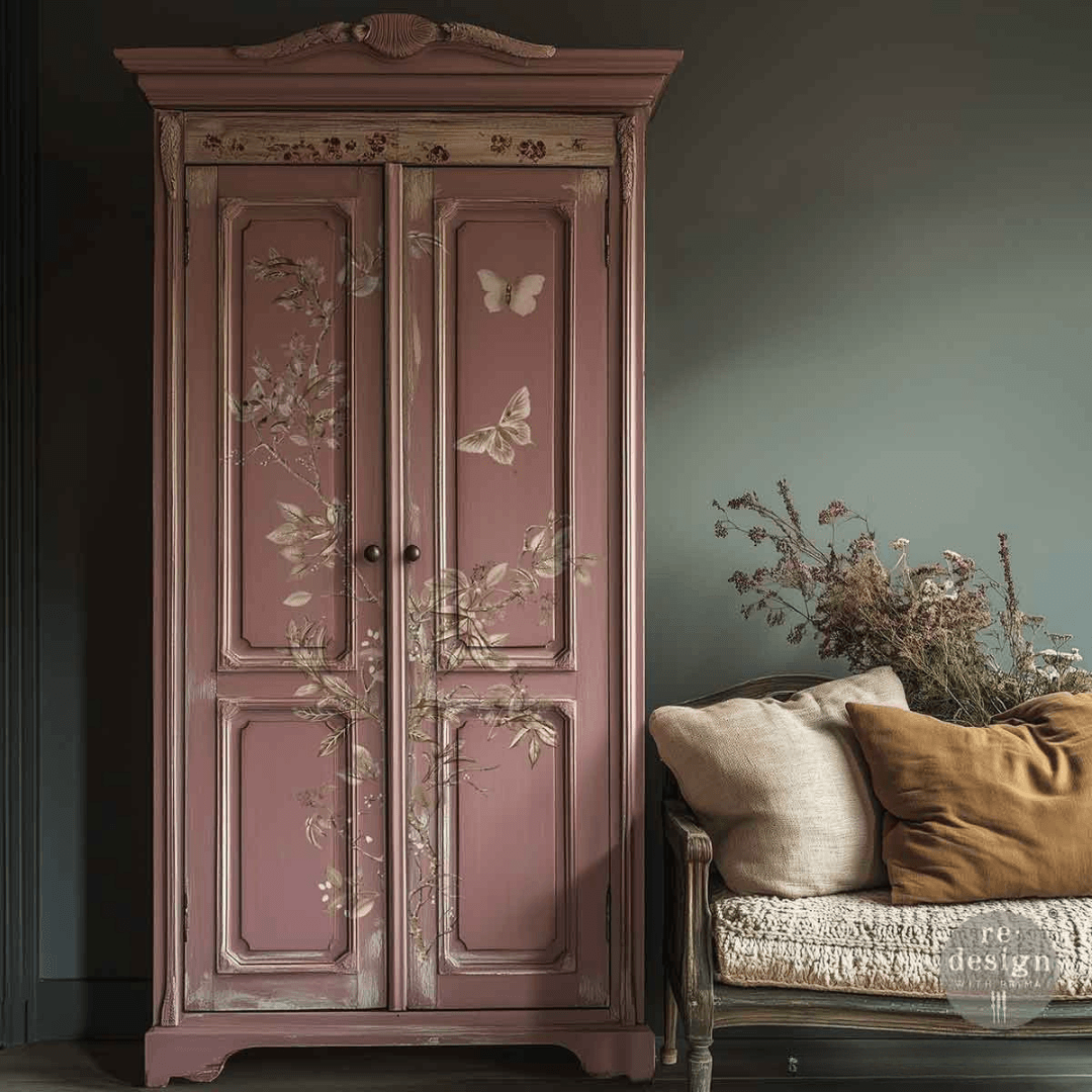 A tall, distressed pink wooden armoire with hand-painted butterfly and floral motifs, positioned next to a vintage-style bench with neutral and mustard-colored cushions against a dark gray wall.
