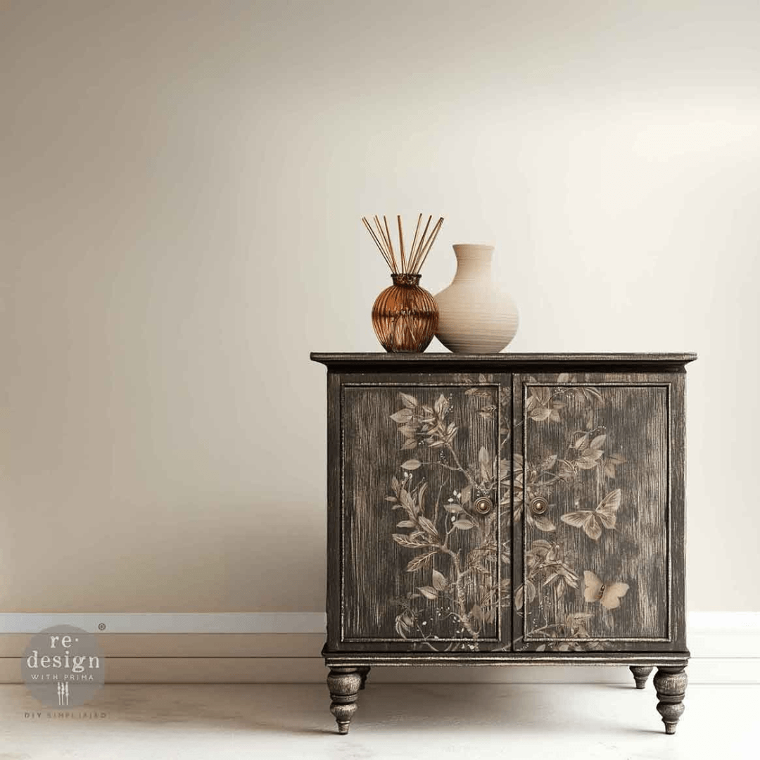 A dark distressed wood cabinet with floral and butterfly motifs on the doors, styled with a reed diffuser and a ceramic vase on top, set against a neutral beige wall.