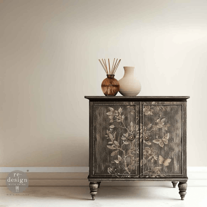 A dark distressed wood cabinet with floral and butterfly motifs on the doors, styled with a reed diffuser and a ceramic vase on top, set against a neutral beige wall.