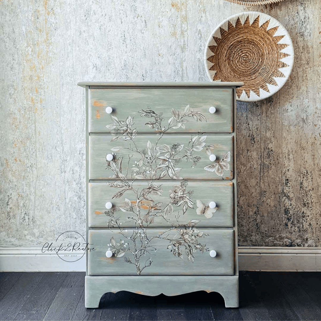 A four-drawer chest of drawers in a distressed sage green finish, decorated with hand-painted floral and butterfly motifs across the front. It features small white knobs and stands on a dark wood floor against a textured, rustic wall with a woven wall hanging.