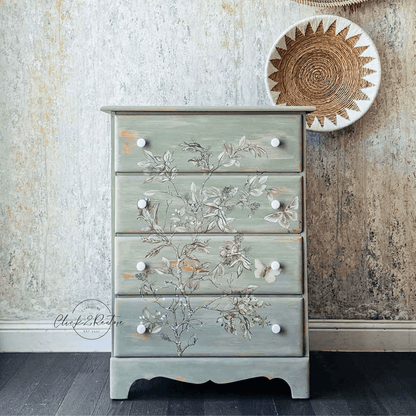 A four-drawer chest of drawers in a distressed sage green finish, decorated with hand-painted floral and butterfly motifs across the front. It features small white knobs and stands on a dark wood floor against a textured, rustic wall with a woven wall hanging.