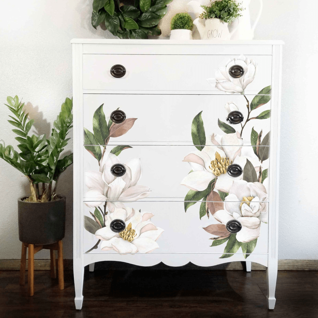 A white four-drawer dresser with scalloped legs and black round knobs, decorated with large painted white flowers, green leaves, and soft brown accents. The dresser stands on a dark wood floor against a white wall, with potted plants nearby.