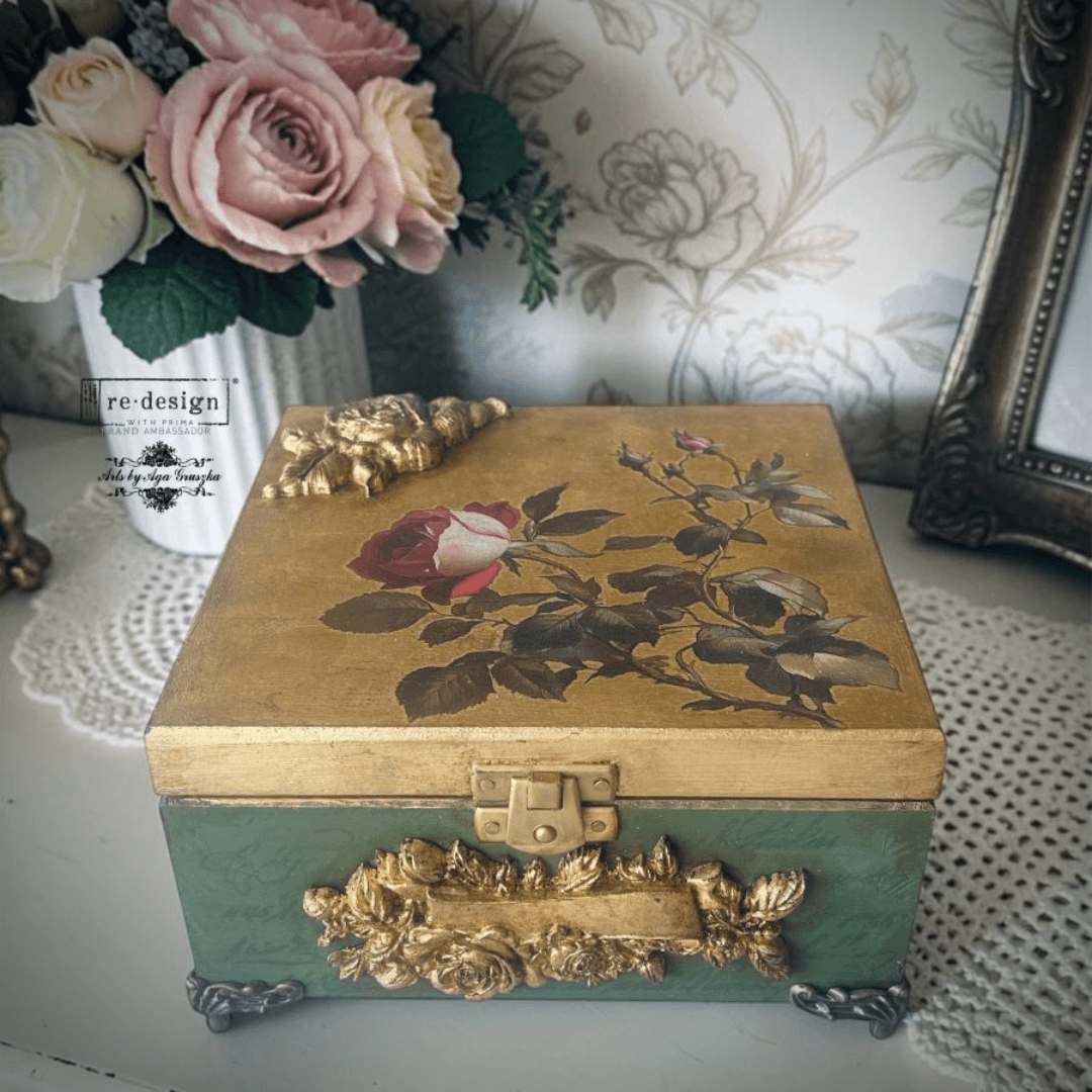 A vintage-style decorative storage box with a gold top featuring a painted rose design, a muted green front, and ornate gold metal accents, styled on a lace doily with flowers and wallpaper in the background.